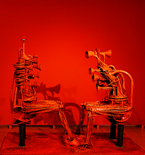 Deux personnages anthropomorphes - entièrement construits à partir de matériaux métaliques de récupération et de fers forgés - sont assis l'un en face de l'autre. Fond monochome rouge en surimpression.