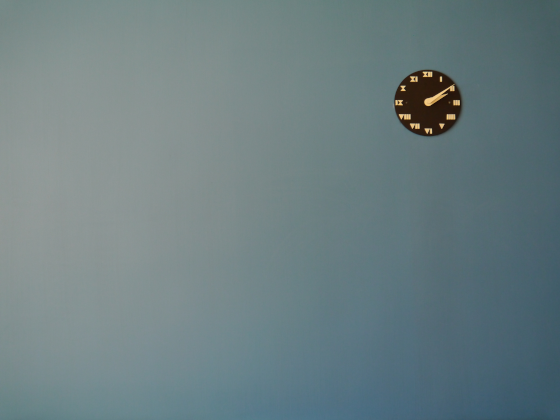 Un mur uni de couleur bleu gris avec juste une pendule placée en haut et à droite de la photo.